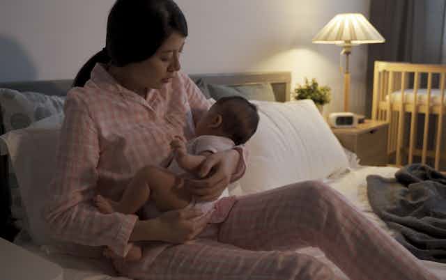 A woman in pajamas on a bed holding a baby with a crib in the background