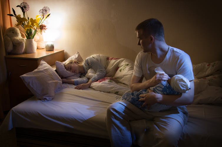 A man is feeding a bottle to a baby in bed while a woman sleeps behind him.