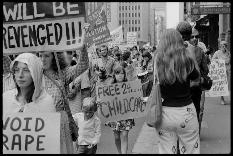 A child in a protest holds a sign reading 'free 24 hour childcare'