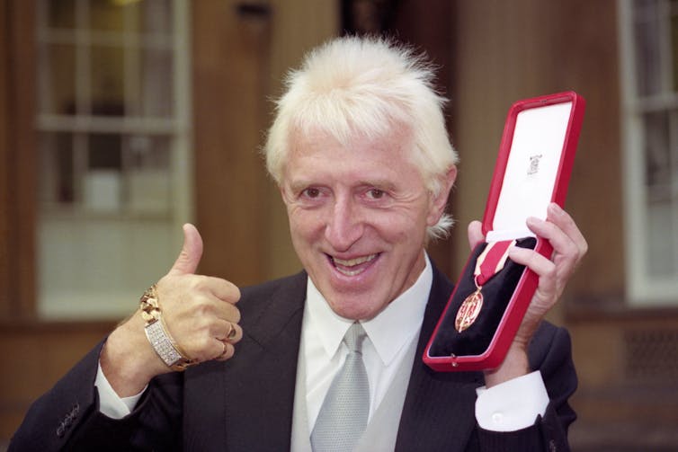 Jimmy Savile in a suit and tie holds up a medal in one hand and gives a thumbs up, with a bling bracelet, with the other.