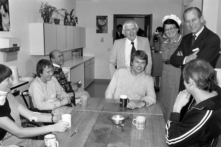 A black and white photograph of hospital patients and staff, Prince Philip and Jimmy Savile sitting around a table, drinking tea.