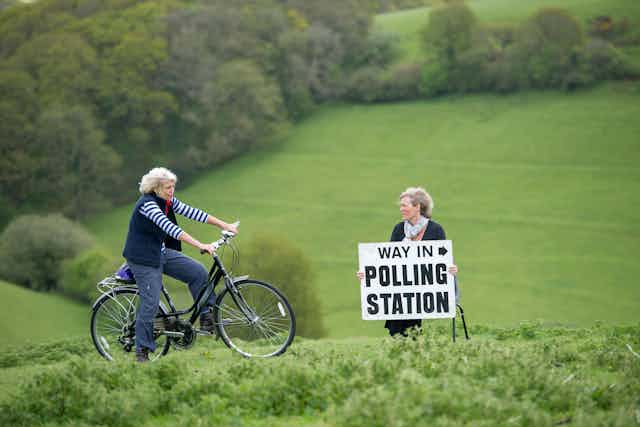 A woman rides her bike past another woman sitting on a chair in the middle of a field holding a sign reading 'way in, polling station'.