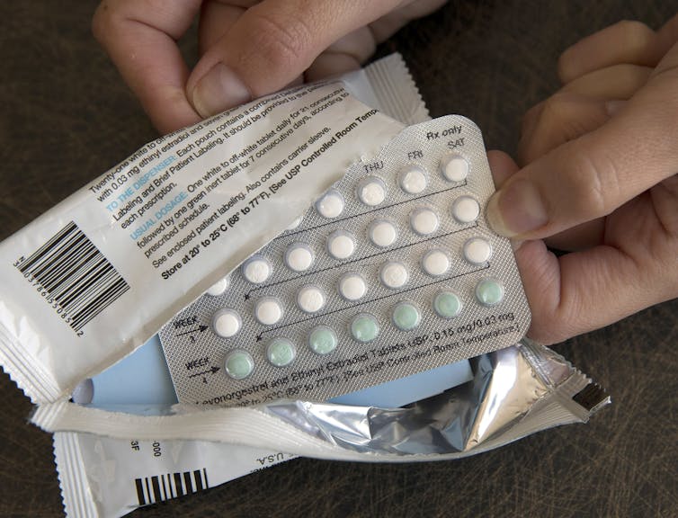 two hands hold a plastic package that's been ripped open showing a container containing many little white and green pills