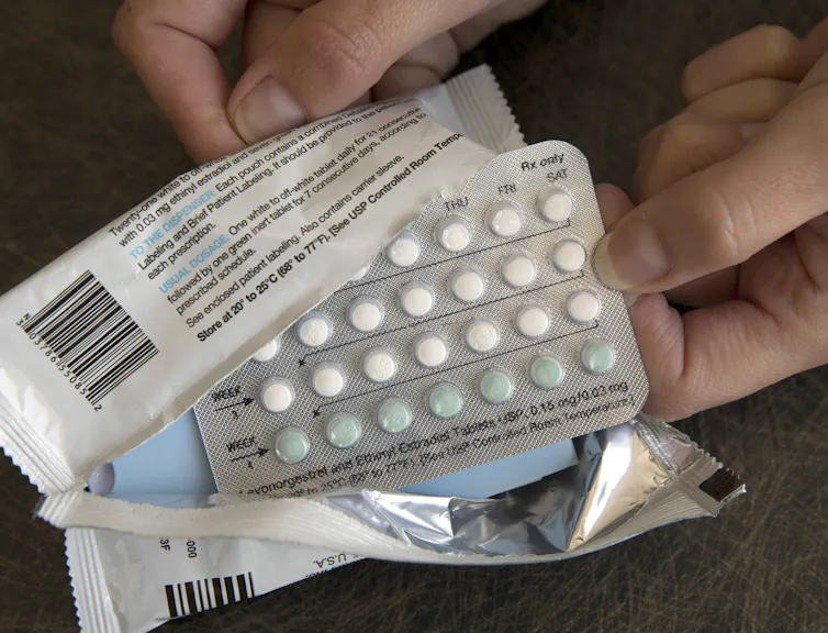 two hands hold a plastic package that's been ripped open showing a container containing many little white and green pills