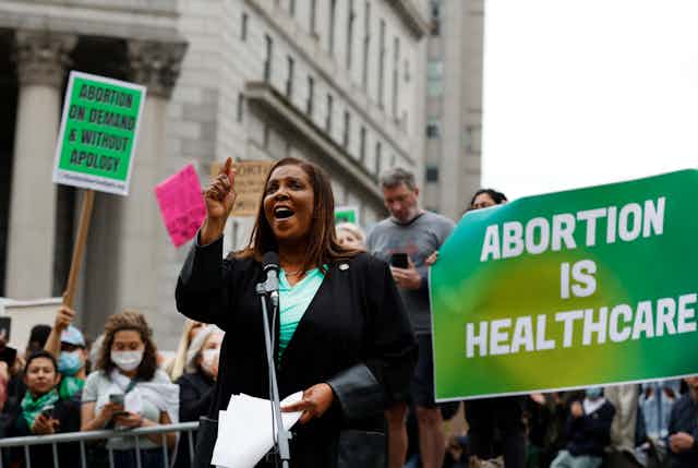 A black woman wearing a dark jacket over a green shirt raises her right hand in a gesture and holds papers in her right as she speaks at a rally at a rally in front of a large stone building and a crowd of protesters. some carrying signs, one reading 'abortion is healthcare'