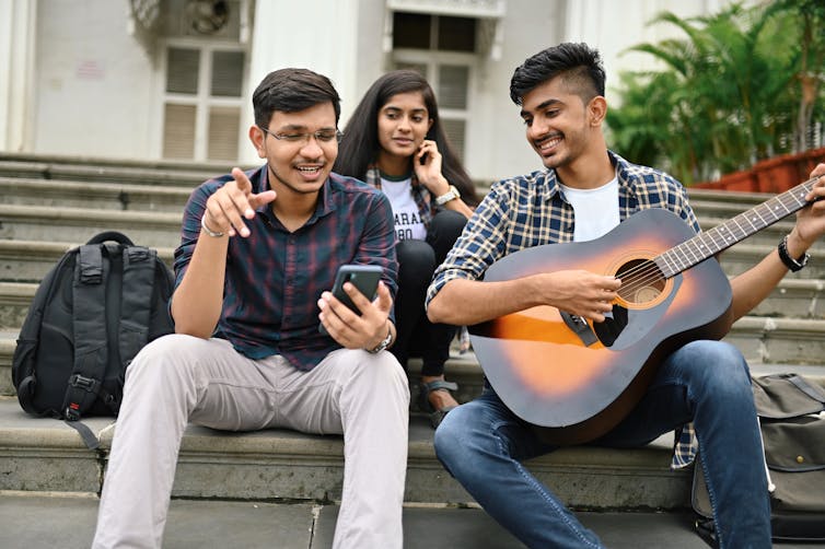 A student plays the guitar on the steps of a building while his friends listen.