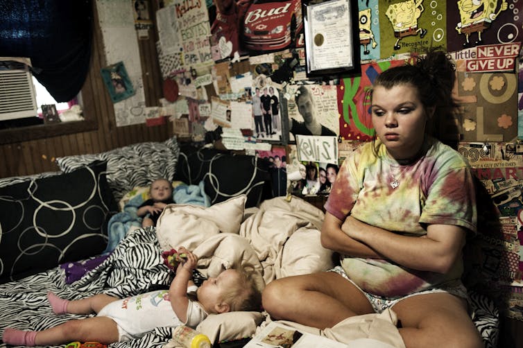 A teenage girl sits in a bedroom with posters on the wall, arms crossed.  Around her on the bed lie two small children.