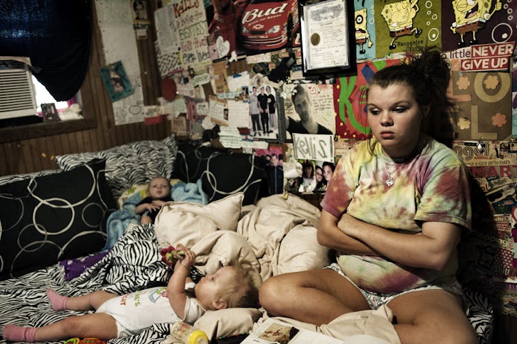 A teenage girl sits in a bedroom with posters on the wall, with her arms crossed. Two young children lie on the bed around her.