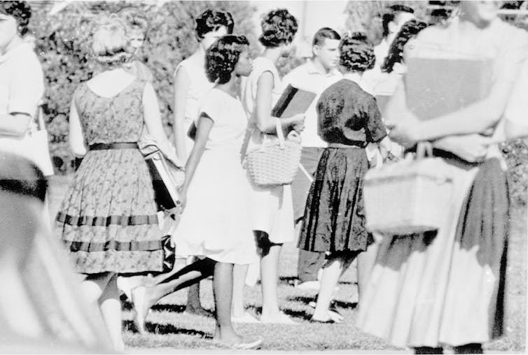 A young Black girl in a white dress walks through a crowd of white people