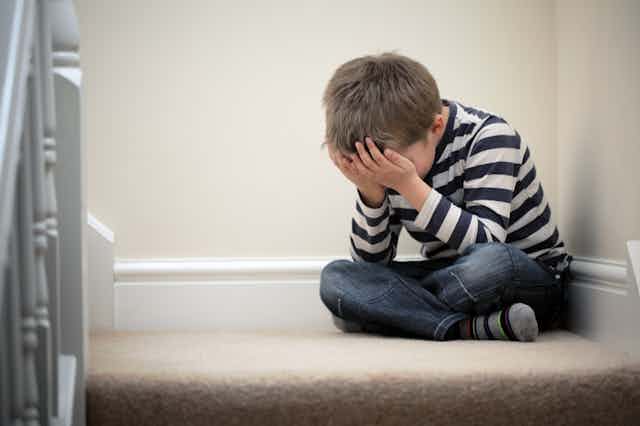A young boy, his hands covering his face, sits on a staircase in despair.