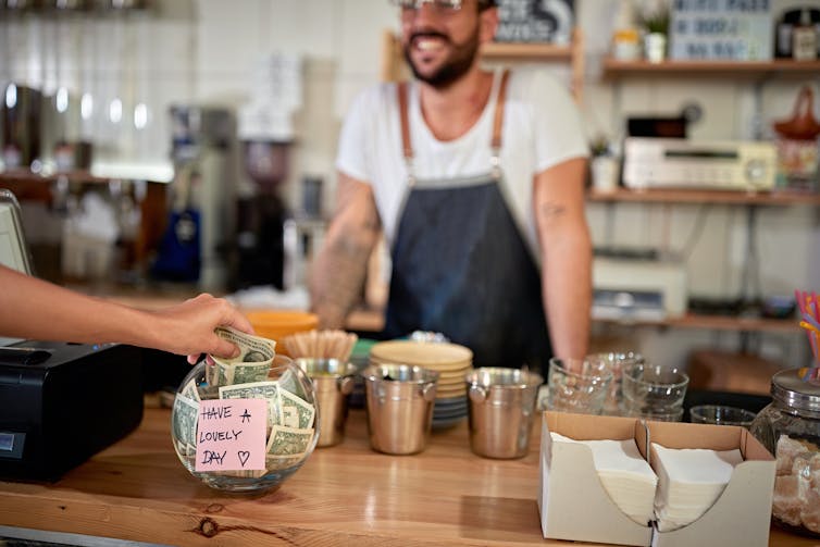 A barista standing behind a counter with a tip jar on it. A customer is putting money in the jar.