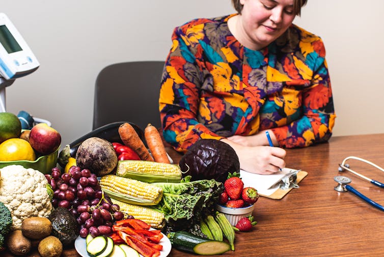 A woman writing on a clipboard at a table with a stethoscope and a big selection of fruits and vegetables on it