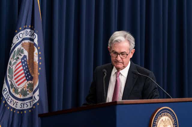 A white man with gray hair looks down as he stands near a podium with a seal on it in front of a blue curtain and a flag that says 'federal reserve system' on it
