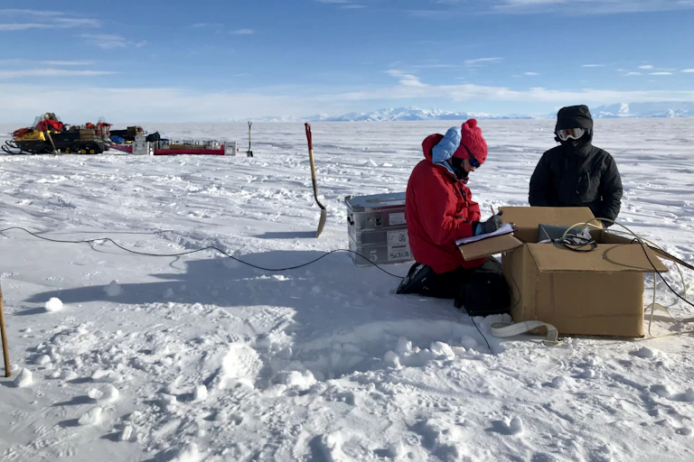 Two people in thick snowsuits kneel on the ice next to a box with gear. Snowmobiles, shovels and mountains are in the background.