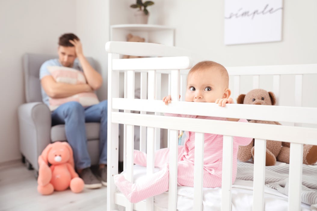 Baby in cot with father in background clutching cushion, holding head