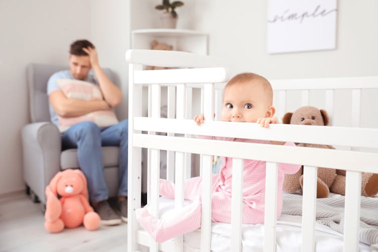 Baby in cot with father in background clutching cushion, holding head