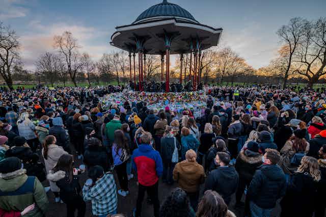 People stand vigil around a bandstand at dusk in a park.