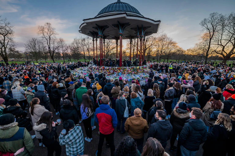 People stand vigil around a bandstand at dusk in a park.