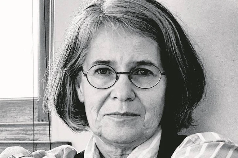 A black and white image of a woman with greying hair in a bob, round spectacles on, looking directly into camera, face open.