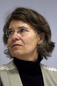 An earnest woman looks over her shoulder, round glasses on.