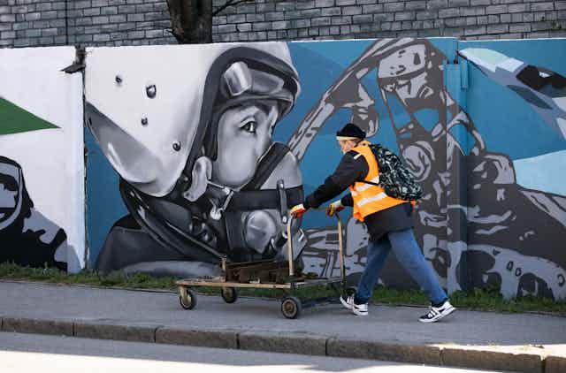 A man is pushing a cart down the street in the Russian enclave of Kaliningrad. Graffiti on the wall with a portrait of a man in an aviation helmet, April 2022.