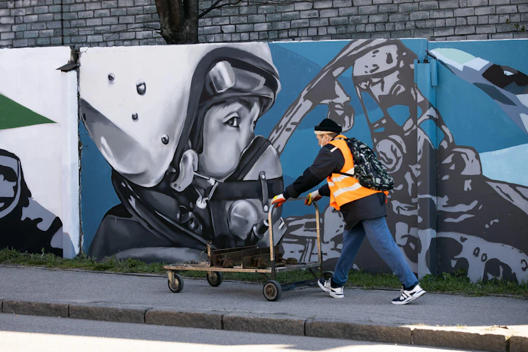 A man is pushing a cart down the street in the Russian enclave of Kaliningrad. Graffiti on the wall with a portrait of a man in an aviation helmet, April 2022.