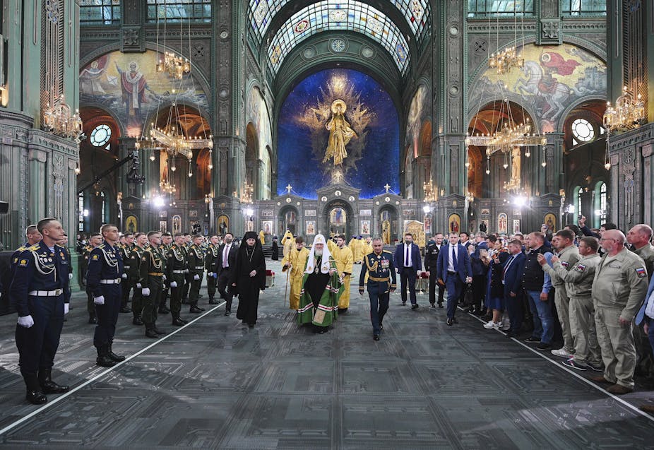 An Orthodox priest in robes walks through a grand cathedral, as men in military uniforms stand on the sides.