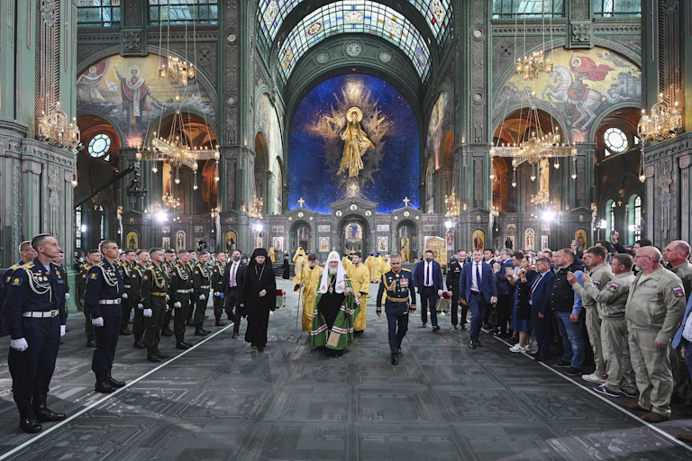 An Orthodox priest in robes walks through a grand cathedral, as men in military uniforms stand on the sides.