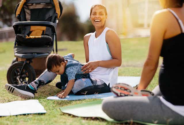 A woman in fitness clothes sitting on a mat on grass with a child in her lap and a stroller beside her, and another woman in fitness clothes seen from behind