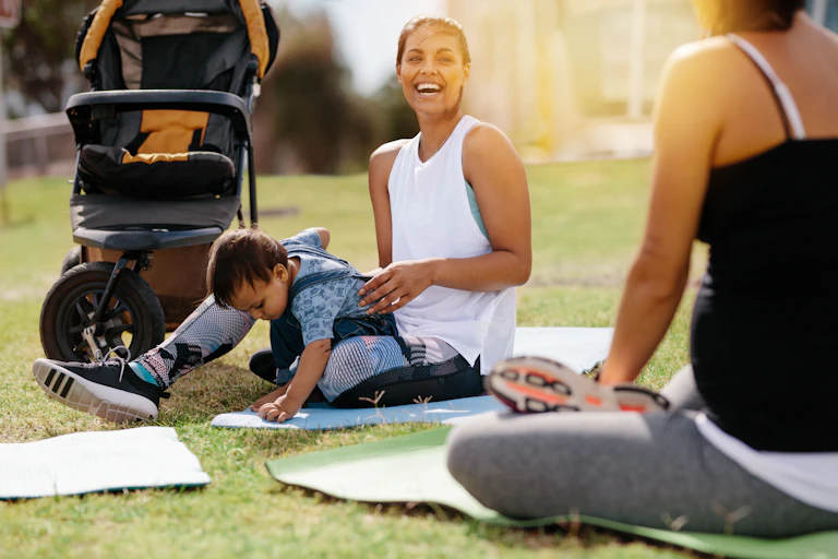 A woman in fitness clothes sitting on a mat on grass with a child in her lap and a stroller beside her, and another woman in fitness clothes seen from behind