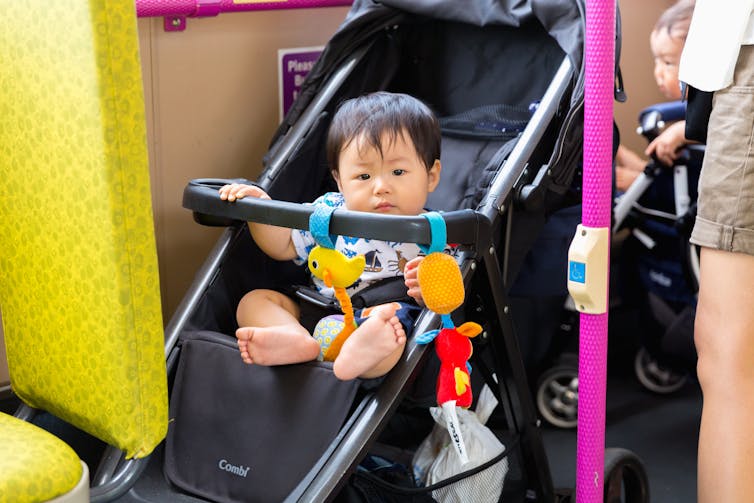 A baby in a stroller behind a yellow bus seat and a pink pole with a button on it