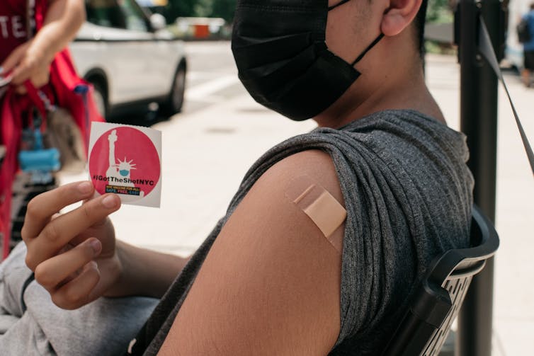 person holds up a vaccination sticker next to arm with bandaid