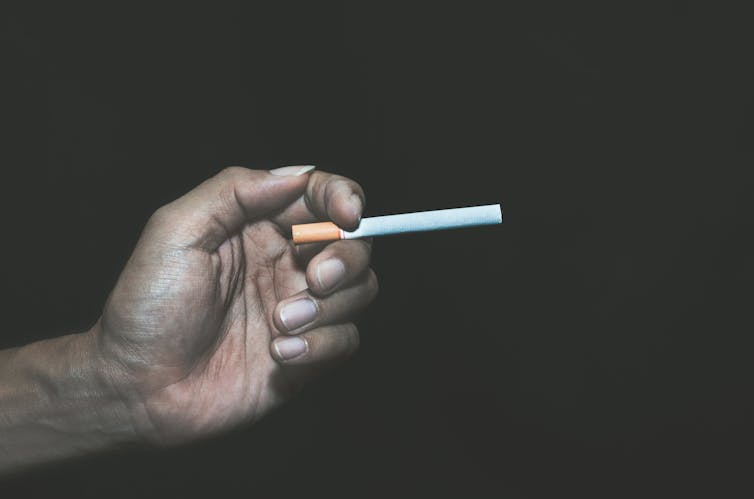 Against a black background, an unidentified hand with dark skin, dirty nails and a generally unhealthy look holds an unlit cigarette between to fingers.