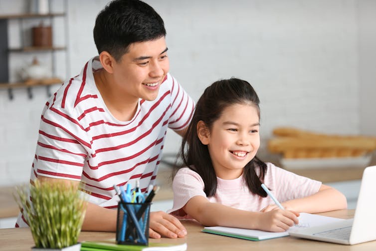 a girl sitting at a table, her father behind her helping with homework