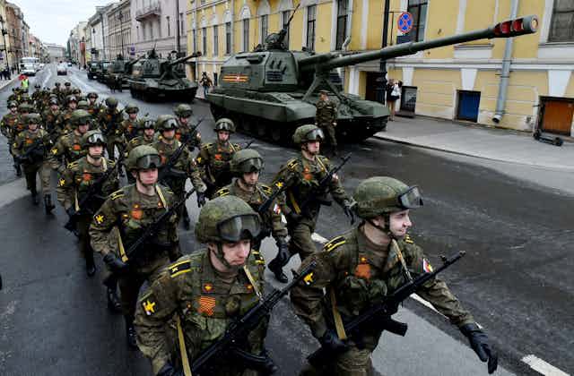 Two rows of Russian soldiers in uniform march down an empty street with tanks on the side 
