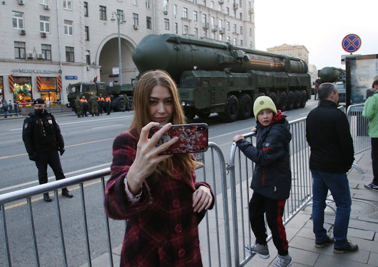 A woman smiles and takes a selfie in front of a Russian tank on a guarded street in Saint Petersburg, blocked off with barricades