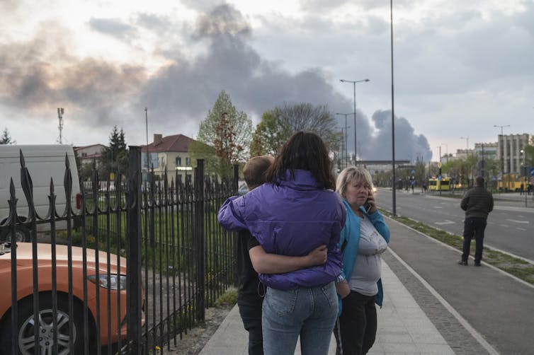 Two people hug and one woman talks on her cellphone on a street. In the background are large billowing clouds of smoke from missile strikes.