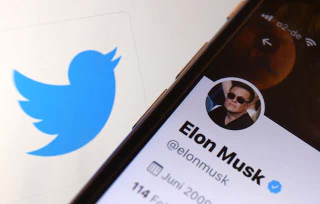 a smart phone with its screen displaying Elon Musk's Twitter account on a surface containing a blue cartoon silhouette of a bird