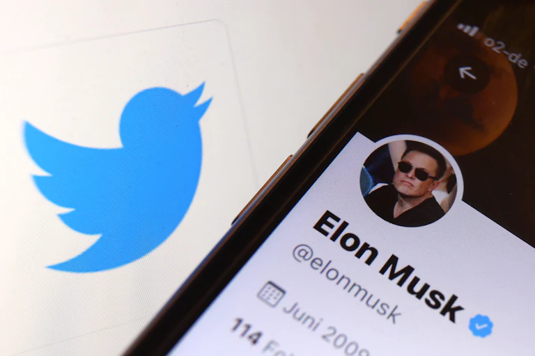a smart phone with its screen displaying Elon Musk's Twitter account on a surface containing a blue cartoon silhouette of a bird