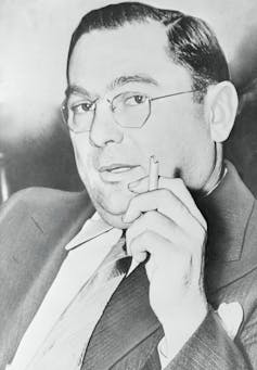 Man with glasses poses holding a cigarette.