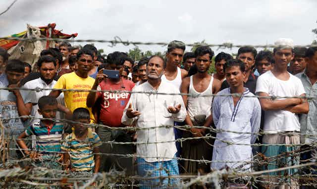 A group of Rohingya men and boys standing behind a barbed wire fence.