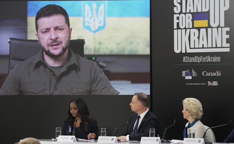 A screen displays a man in a button up and three people sit on a panel in front.