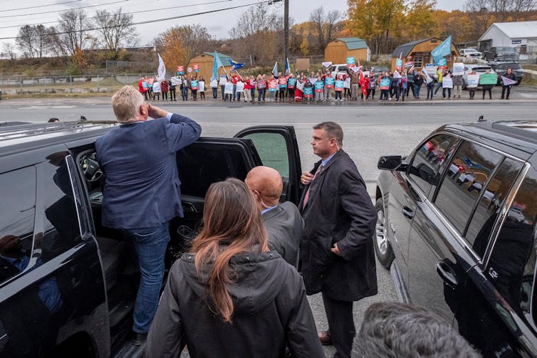 A man gets into a car with as a line of protesters stand in a row across the street holding signs.