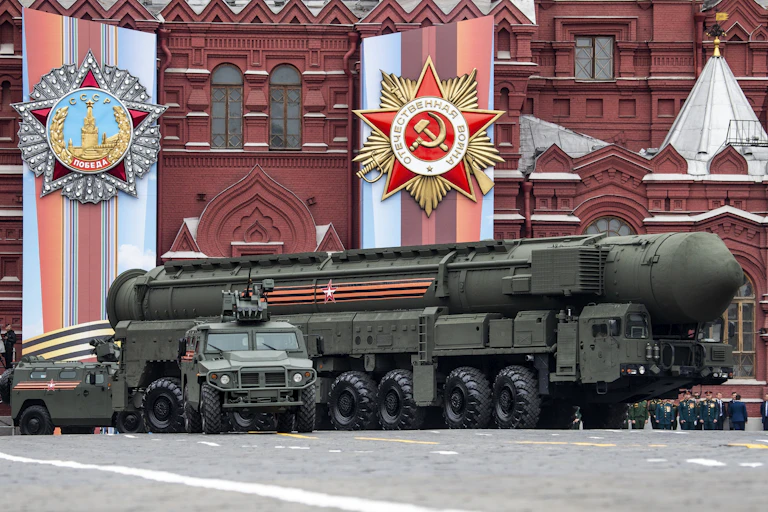 Military personnel are seen in front of a giant missile with the decorative red facades of buildings behind them.