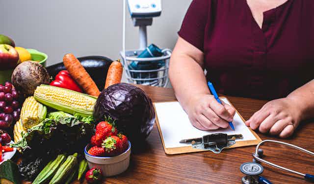Fresh vegetables and fruit on a desk beside a clipboard with a person's hand writing on it and a stethoscope