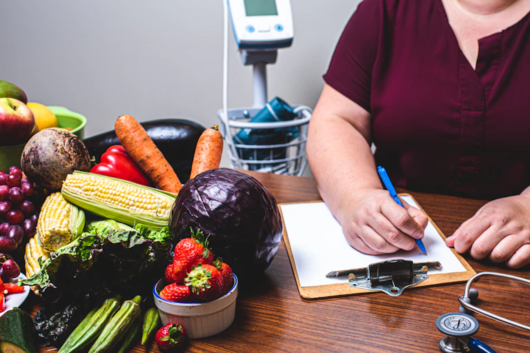 Fresh vegetables and fruit on a desk beside a clipboard with a person's hand writing on it and a stethoscope