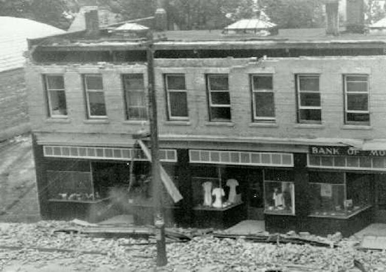 A black and white photo of a two-storey building with bricks strewn in the street.