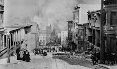 A black and white photo of people standing the street, watching smoke billow from the city below.