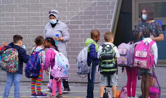 Teachers in face masks seen greeting students in a schoolyard getting ready to go inside.