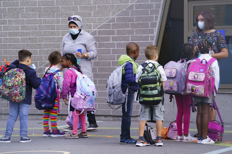 Teachers in face masks seen greeting students in a schoolyard getting ready to go inside.